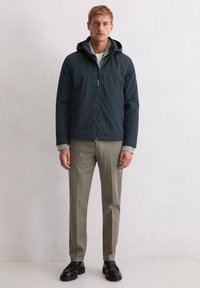 Kurtka z kapturem w ciemnym niebieskim kolorze, wykonana z lekkiego materiału, zapinana na zamek błyskawiczny, z bocznymi kieszeniami. Połączona z beżowymi spodniami i czarnymi butami.