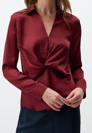Femme portant un chemisier en satin bordeaux à manches longues avec un détail torsadé à l'avant, associé à une jupe foncée, mains se touchant doucement.