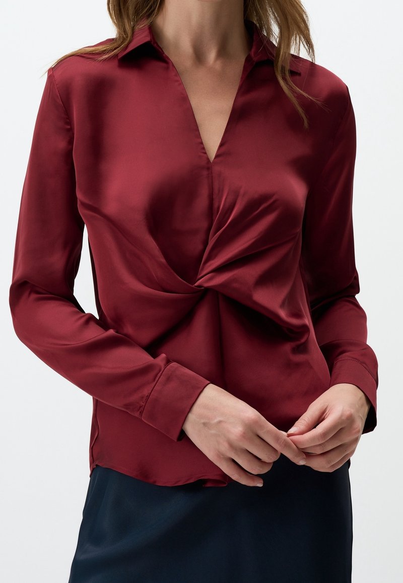 Femme portant un chemisier en satin bordeaux à manches longues avec un détail torsadé à l'avant, associé à une jupe foncée, mains se touchant doucement.