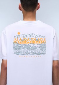 Camiseta blanca de algodón con un diseño gráfico en la parte trasera que presenta montañas, un sol y "NAPAPIJRI" en colores naranja y verde.