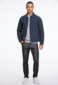 Marineblauwe jas met knopen en een voorzak, gelaagd over een grijze sweatshirt, gecombineerd met donkere jeans en witte sneakers.