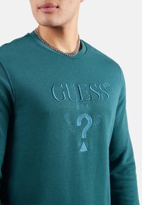 Sudadera de manga larga en color verde azulado con el logotipo bordado de "GUESS" y diseño de signo de interrogación. Presenta un escote redondo y una textura suave y lisa.