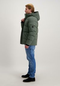 Groene pufferjack met capuchon, voorzien van een zijzak en een rond logo-embleem. Draagt met een blauwe spijkerbroek en zwarte enkellaarzen.