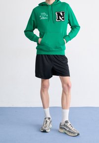 Grön hoodie med en stor svart "N"-logotyp och vit detaljering, tillsammans med svarta shorts och grå sneakers. Har en framficka och en huva.