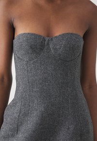 Robe grise sans bretelles en chevrons avec bonnets structurés, tissu texturé et silhouette ajustée. Des coutures tailleur soulignent la forme.