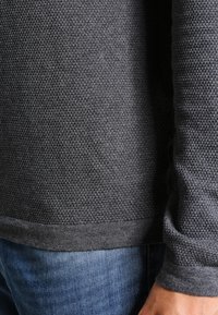 Tmavě šedý pletený svetr s texturovaným vzorem, s žebrovaným lemem a manžetami, kombinovaný s modrými džínami.