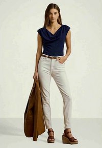 Γυναίκα με ναυτικό μπλε ανάλαφρη αμάνικη μπλούζα, λευκά παντελόνια ψηλής μέσης, καφέ πλατφόρμες, κρατώντας ένα καφέ σακάκι, standing against a plain background.