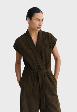 Femme aux cheveux foncés portant une combinaison marron foncé texturée avec ceinture, les mains dans les poches, regardant vers sa droite sur un fond uni clair.