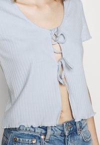 Žena v světle modrém žebrovaném krátkém rukávu, na kterém jsou dvě mašle vpředu, v kombinaci s modrými džínami s vysokým pasem.