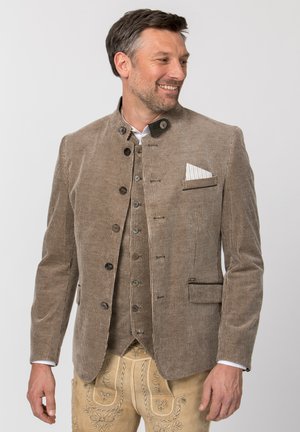 Mann lächelnd mit einer braun gestreiften, geknöpften Jacke, passender Weste, weißem Hemd und beigen bestickten traditionellen Hosen.