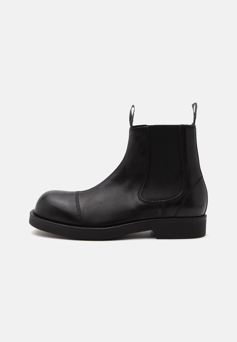Bottine Chelsea en cuir noir à bout arrondi, panneaux latéraux élastiques, languettes à l’avant et à l’arrière, semelle plate, vue de côté sur fond blanc.