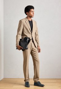 Hombre con traje beige y camisa negra sosteniendo un gran bolsa de cuero texturizado negro, de pie sobre un suelo de madera contra una pared lisa de color claro.