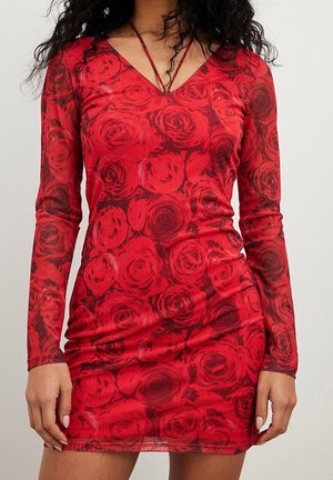 Femme portant une robe rouge moulante en mesh à manches longues avec un motif de roses et un décolleté en V profond.
