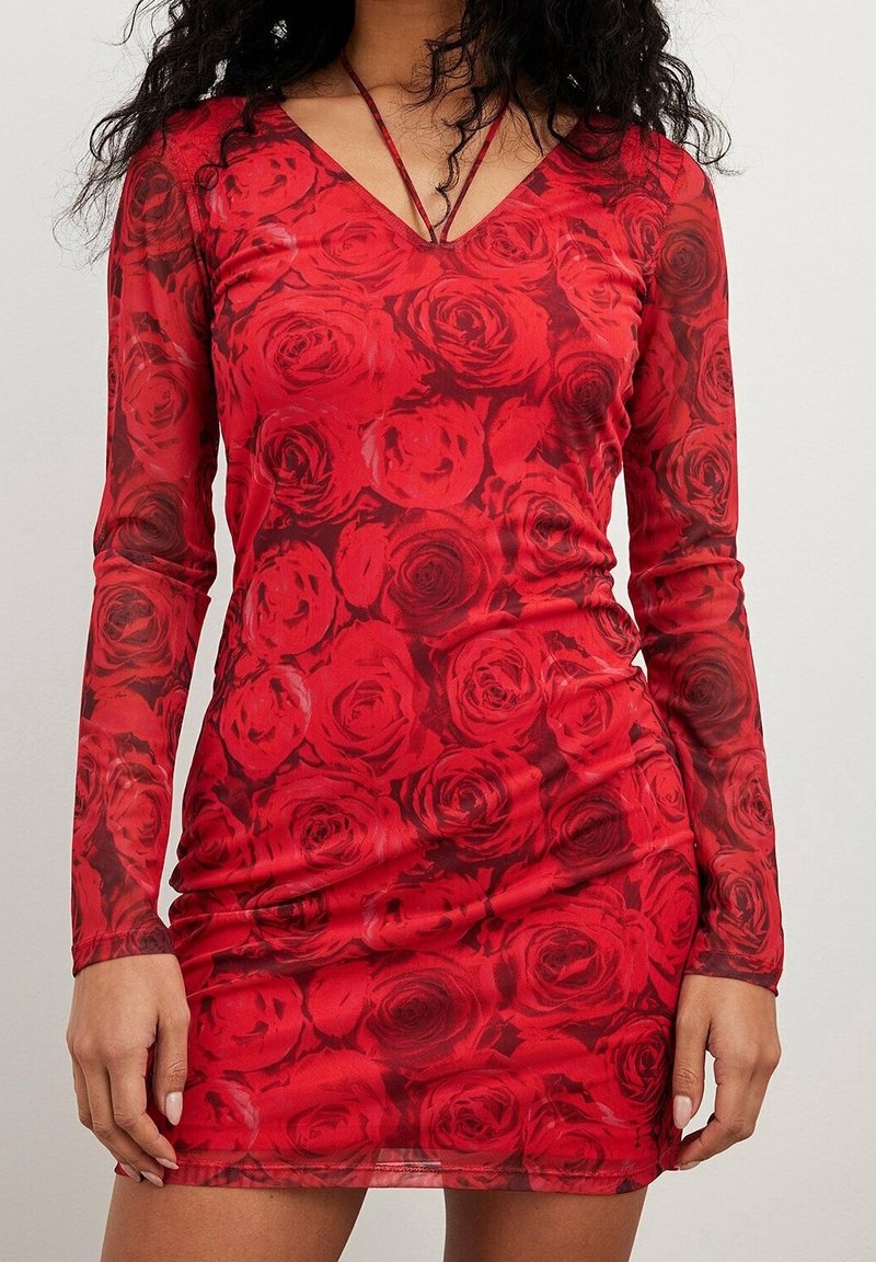 Femme portant une robe rouge moulante en mesh à manches longues avec un motif de roses et un décolleté en V profond.