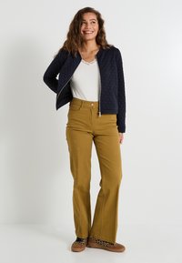 Femme avec des cheveux ondulés portant une veste matelassée navy, un haut blanc avec des dentelles, un pantalon large moutarde et des chaussures à imprimé léopard, debout en souriant.