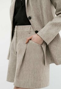 Shorts beige in lino su misura abbinati a un blazer e una maglia nera, con pieghe laterali e una mano appoggiata nella tasca.