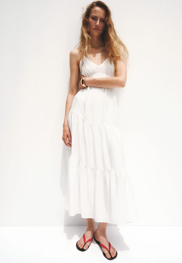 Jeune femme aux longs cheveux ondulés porte une robe maxi blanche sans manches et des tongs rouges, debout les bras croisés sur un fond blanc.