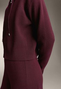 Detailný záber osoby v zladenom vínovom pletenom mikinách s kapucňou a nohaviciach so zipsom a šnúrkami.