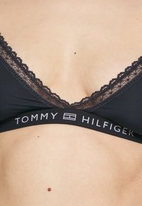 Musta bralette pitsireunalla kaula-aukolla, jossa on merkkijoustonauha, jossa lukee "TOMMY HILFIGER" valkoisella tekstillä.