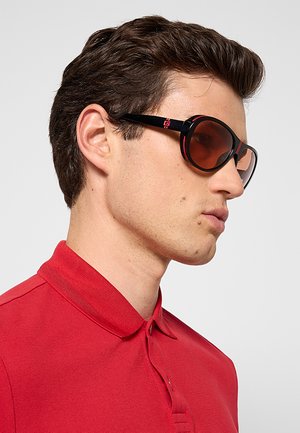Uomo con occhiali da sole neri e rossi e polo rossa, di profilo rivolto verso destra su uno sfondo chiaro uniforme.