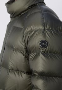 Chaqueta acolchada verde oliva hecha de tejido metálico, con secciones acolchadas, un cuello alto y un parche redondo negro del logo en el hombro.