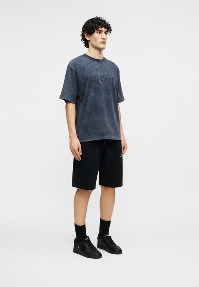 Νεαρός άνδρας στέκεται, φορώντας ένα ξεβαμμένο μαύρο t-shirt Off-White, μαύρες βερμούδες, μαύρες κάλτσες και μαύρα αθλητικά παπούτσια σε απλό λευκό φόντο.