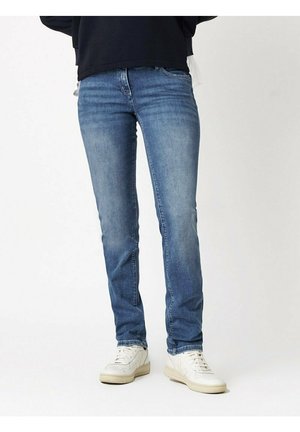 Osoba ubrana w wyblakłe niebieskie obcisłe dżinsy i białe sportowe buty, stojąca na tle jednolitego jasnego tła.