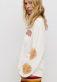 Cremefarbene Sweatshirt mit langen Ärmeln und roten Schriftzügen, mit Sonnenmotiven auf den Ärmeln. Kombiniert mit einem mehrfarbigen Strickrock, der auffällige Muster präsentiert.