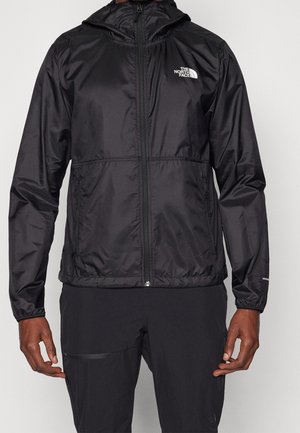 Mężczyzna noszący czarną, zapinaną na zamek kurtkę z kapturem North Face oraz czarne spodnie, stojący z ramionami swobodnie opuszczonymi wzdłuż boków.