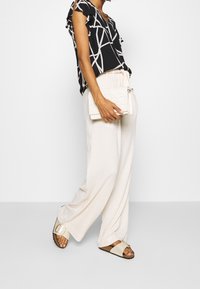 Černý top s bílým geometrickým vzorem, krémové kalhoty s širokými nohavicemi a béžové sandály se zlatou sponou. Drží strukturovanou bílou kabelku.