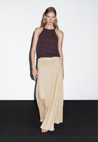 Donna che cammina su pavimento nero contro parete bianca, indossando un top marrone scuro senza maniche e pantaloni beige a gamba larga, a piedi nudi.