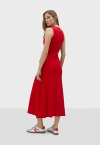 Rode geribbelde gebreide midi-jurk met een aangesloten taille en uitlopende rok, gecombineerd met zilveren en rode sneakers. Effen achtergrond.