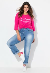 Camisa de manga larga rosa con estampado de "Los Ángeles", anudada en la cintura, combinada con jeans de mezclilla azul claro y zapatillas blancas con acentos rosas.