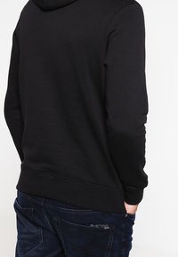 Sudadera negra con una textura suave y bolsillo canguro; cuenta con capucha y puños acanalados. Combinada con jeans de mezclilla oscuros.
