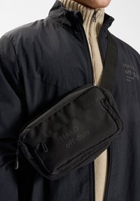 Svart vadderad crossbody-väska med en slät textur och två dragkedjefack. Har ett stämplat logotyp. Bärs över en beige ribbad tröja.