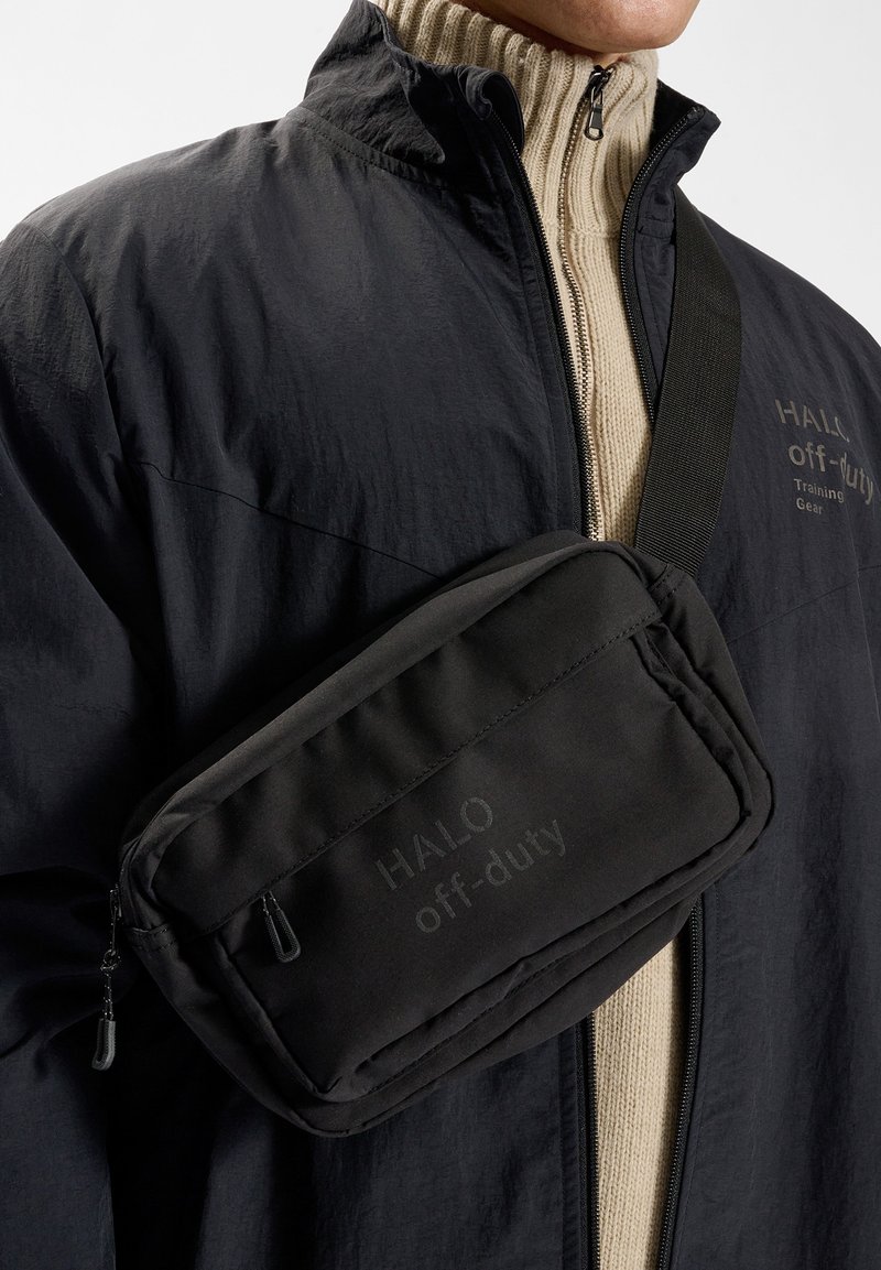 Svart vadderad crossbody-väska med en slät textur och två dragkedjefack. Har ett stämplat logotyp. Bärs över en beige ribbad tröja.