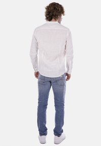 Camicia bianca in lino a maniche lunghe, vista posteriore. Presenta polsini con bottoni e orlo dritto. Indossata con jeans azzurri chiari e sneakers bianche.