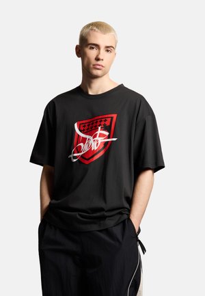 Młoda osoba z krótkimi blond włosami, ubrana w czarny, za duży T-shirt z czerwonym herbem i białym abstrakcyjnym wzorem. Ręce w kieszeniach.