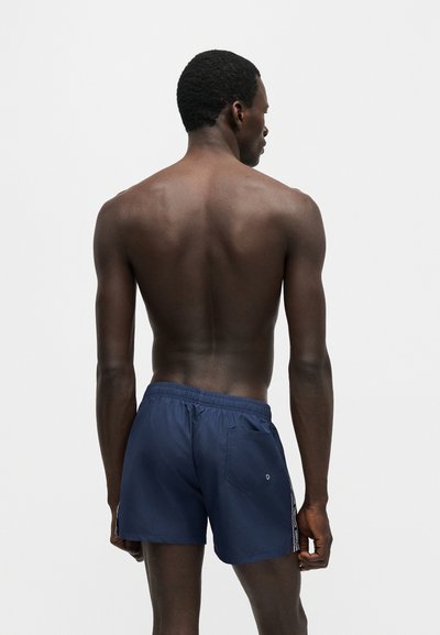 Άνδρας με σκούρο δέρμα και κοντά μαλλιά στέκεται γυρισμένος προς τα πίσω, φορώντας σκούρα μπλε μαγιό, σε απλό φωτεινό φόντο.