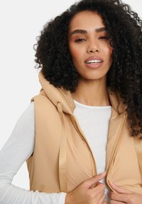 Beige vadderad väst med huva, med en framsida som har dragkedja och kontrasterande guld dragkedjedetaljer. Texturerad tyg med en mjuk yta.