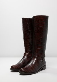 Bottes en cuir marron montant jusqu'au genou avec une texture crocodile, un bout arrondi et une fermeture éclair latérale, dotées d'une sangle décorative à la cheville avec des éléments métalliques.