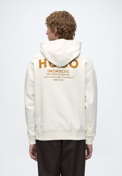 Човек с къдрави коси, носещ бял суичър с кафяв текст "HUGO MCMXCIII Registered Trademark Since 1993" на гърба.