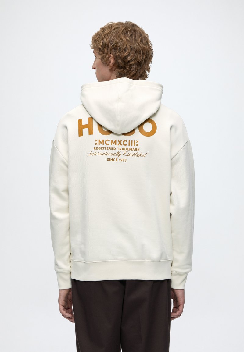 Personne aux cheveux bouclés portant un sweat à capuche blanc avec un texte marron indiquant "HUGO MCMXCIII Registered Trademark Since 1993" dans le dos.