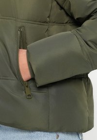 Mână introdusă în buzunarul desfăcut al unei jachete îmblănite verde măsliniu purtată peste blugi de denim albastru deschis.