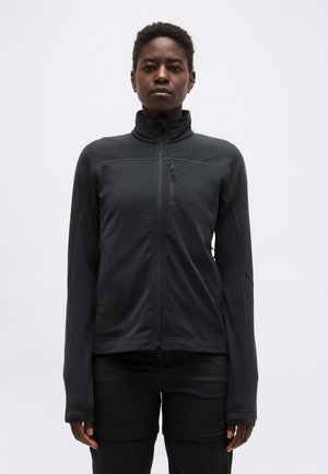 Άτομο που φοράει μαύρο μπουφάν με φερμουάρ, ψηλό γιακά και μακριά μανίκια, στέκεται μπροστά σε απλό ανοιχτό γκρι φόντο.