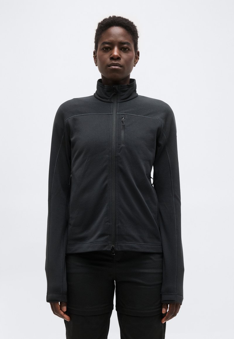 Person wearing a black zip-up jacket with a high collar and long sleeves, standing against a plain light gray background.