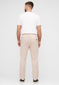 Beige Hosen mit lockerer Passform, elastischem Bund und einer Gesäßtasche, kombiniert mit einem weißen T-Shirt und schwarzen Sneakers. Glatte Textur.
