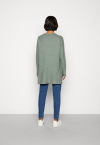 Sweater de mangas longas, verde escuro, com um corte relaxado, rachas laterais e bainha arredondada, combinado com jeans azuis e ténis brancos.