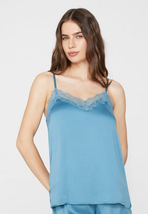 Frau mit langen braunen Haaren, die ein blaues, mit Spitze besetztes Camisole und passende Hosen vor einem einfarbigen hellen Hintergrund trägt.