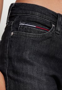 Jean en denim noir au cut classique, présentant une texture subtile, des accessoires en métal argenté, et une petite poche avec des accents rouge, blanc et bleu.